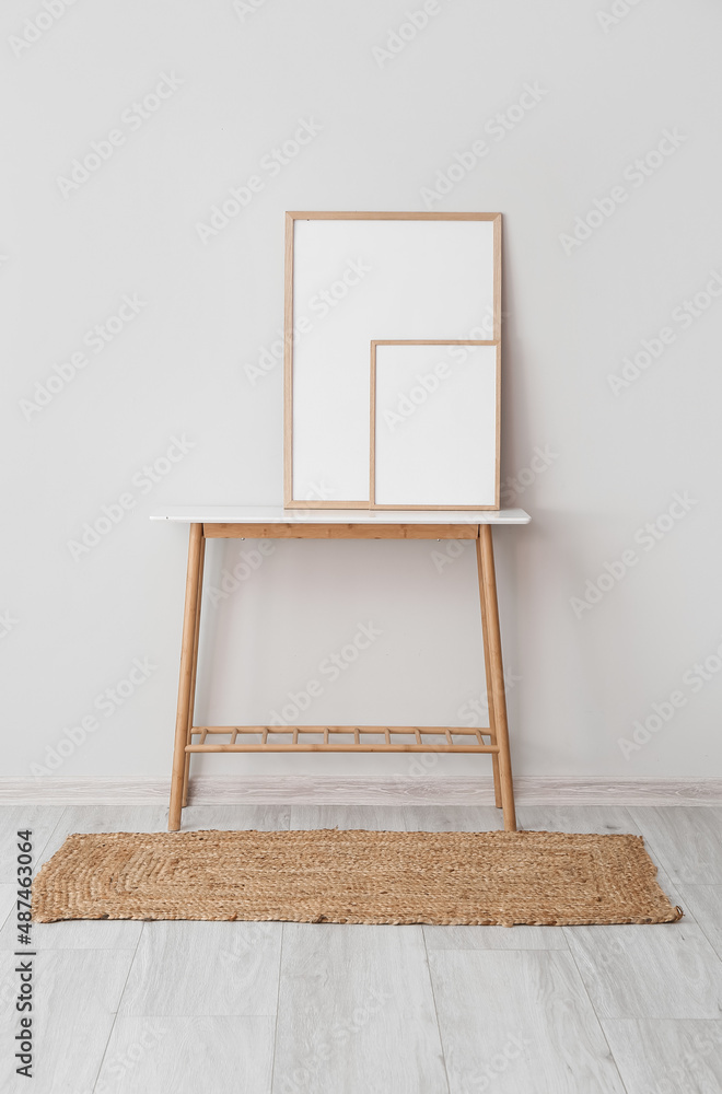 Blank photo frames on table near light wall and wicker carpet on grey wooden floor in room interior