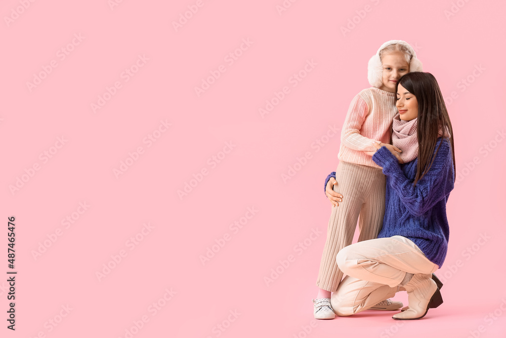 Little girl and her mother in warm sweaters hugging on pink background