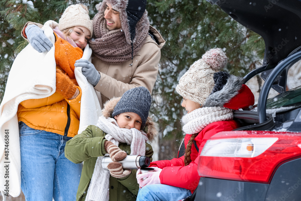 Happy family with thermos drinking hot tea near car in forest on snowy winter day