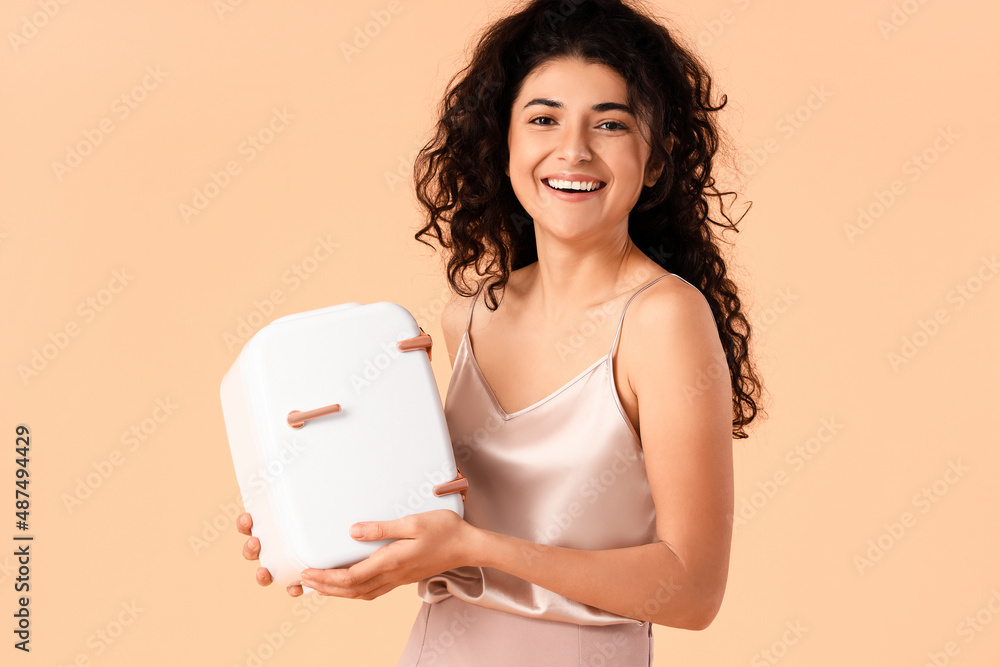 Young smiling woman holding cosmetic refrigerator on color background