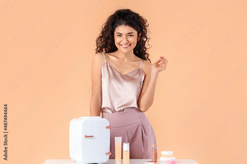 Beautiful smiling woman standing near table with refrigerator and cosmetic products on color backgro