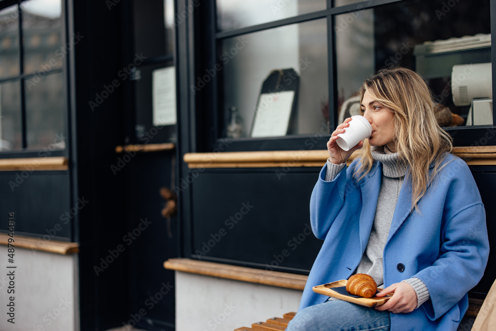 饥饿的高加索妇女穿着蓝色夹克，在吃饭前喝着外卖咖啡。