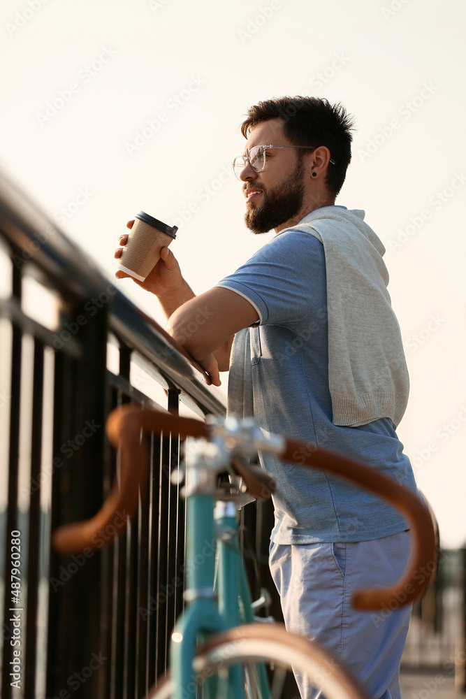 年轻的大胡子男子在路堤上拿着一杯咖啡和自行车