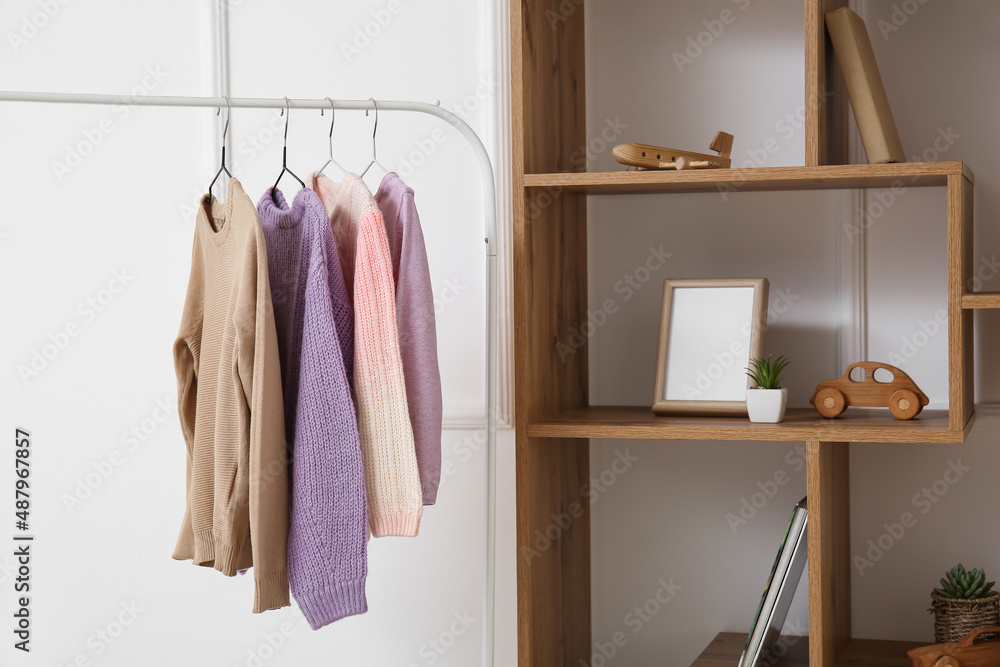 Rack with childrens sweaters and shelf unit near light wall, closeup