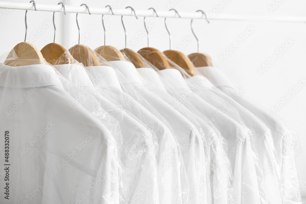 Rack with clean shirts in plastic bags near light wall