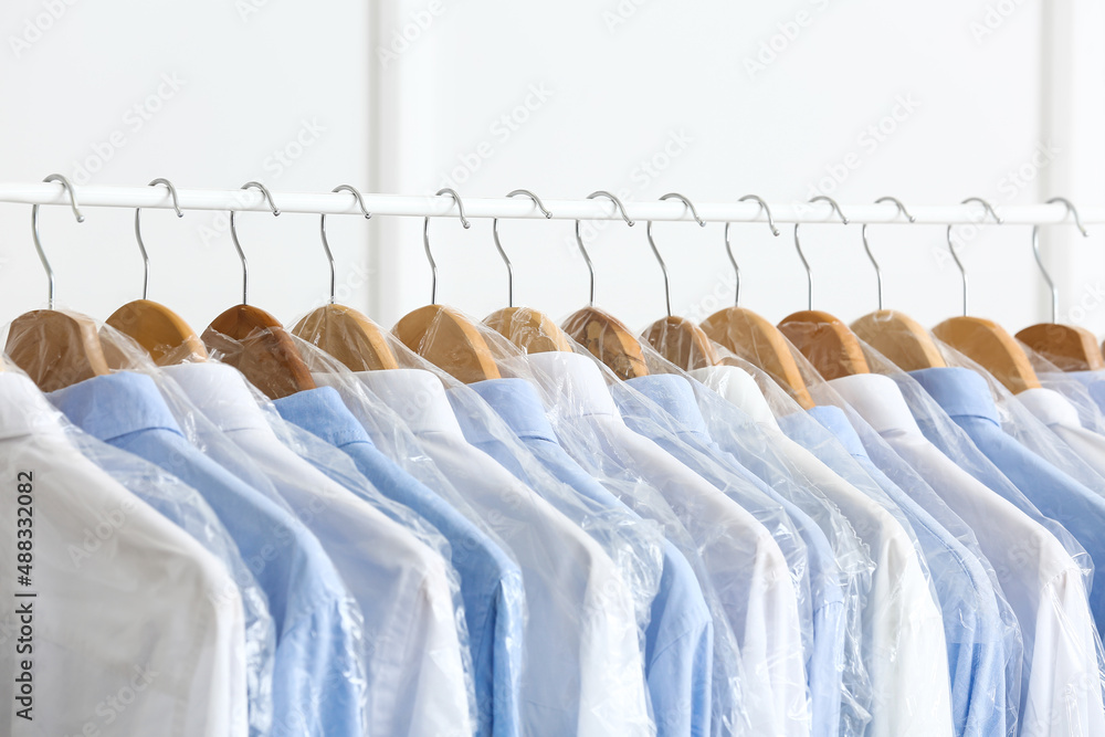 Rack with clean shirts in plastic bags near light wall