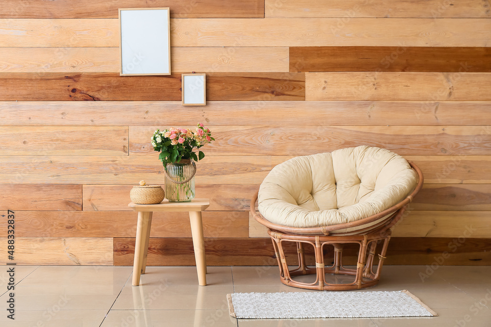 Beautiful roses in vase with basket on table and armchair near wooden wall