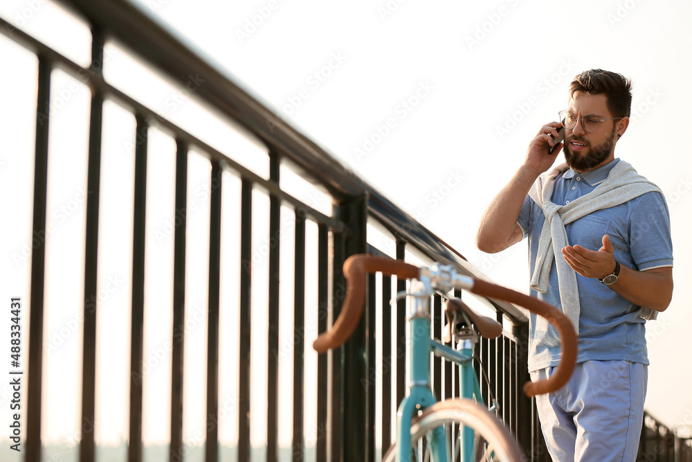 Young bearded man with bicycle talking by mobile phone on embankment