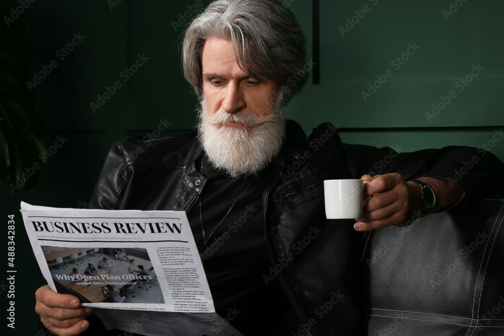 Handsome mature bearded man drinking coffee and reading newspaper at home