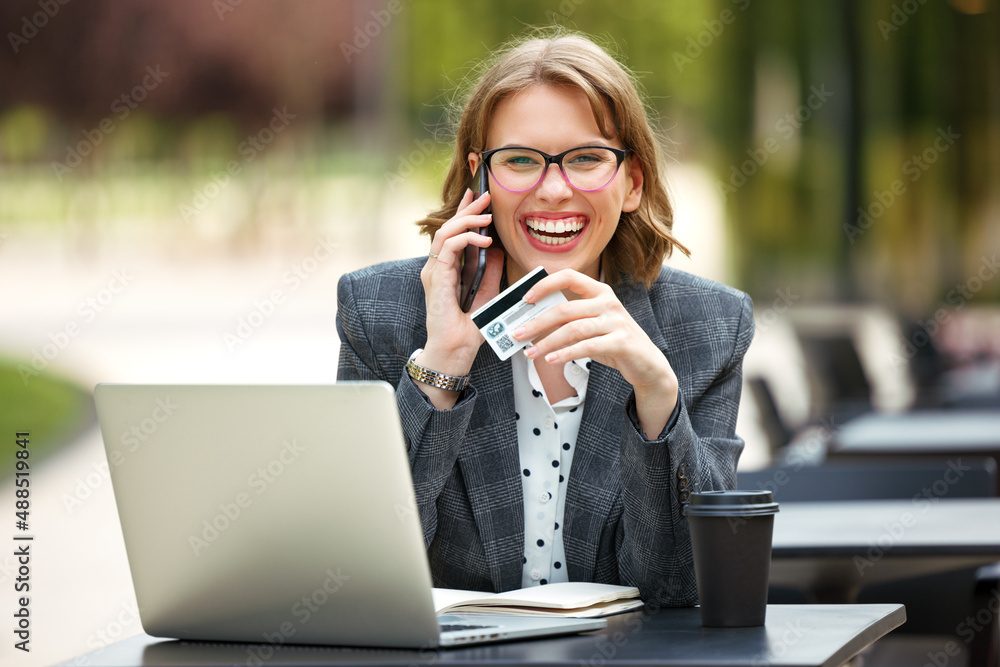 Overjoyed woman with credit card in hand talking on mobile phone while working on laptop outside