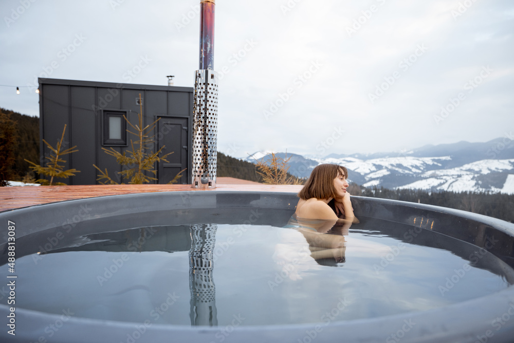 Woman swims in hot bath while resting at small modern house in the mountains on winter. Concept of r