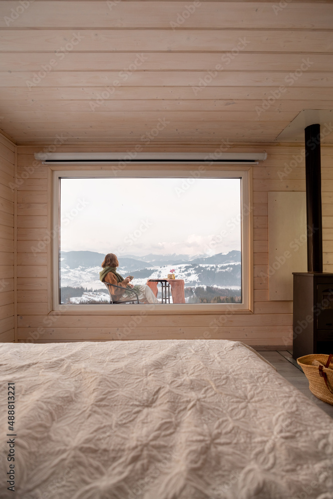 Woman sitting with a drink on terrace of tiny house in the mountains, view through the window from t