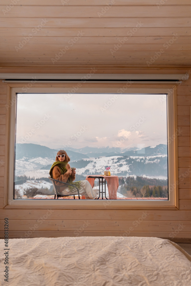 Woman sitting with a drink on terrace of tiny house in the mountains, view through the window from t