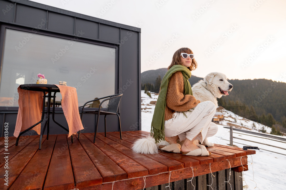 Woman with her dog resting on terrace of tiny house in the mountains on winter time. Concept of smal