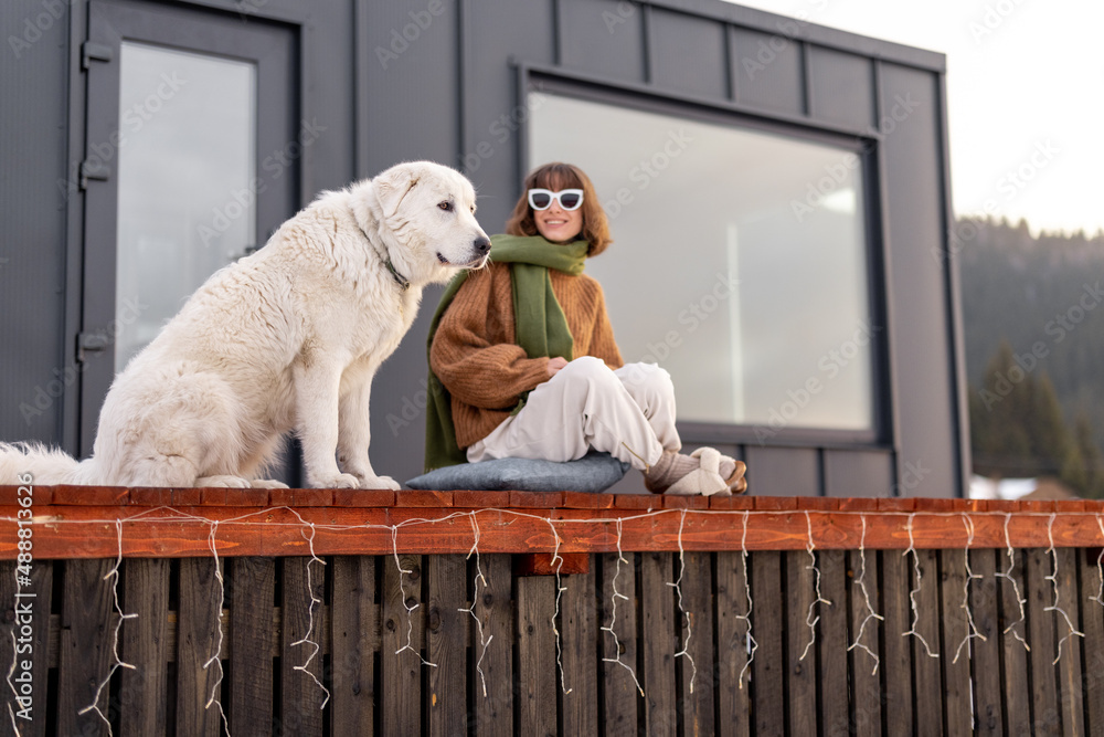 Woman sitting with dog on terrace of tiny house in the mountains. Concept of small modern cabins for