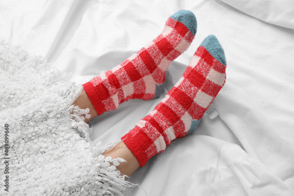 Female legs in knitted socks covered with plaid on bed, closeup. Concept of heating season