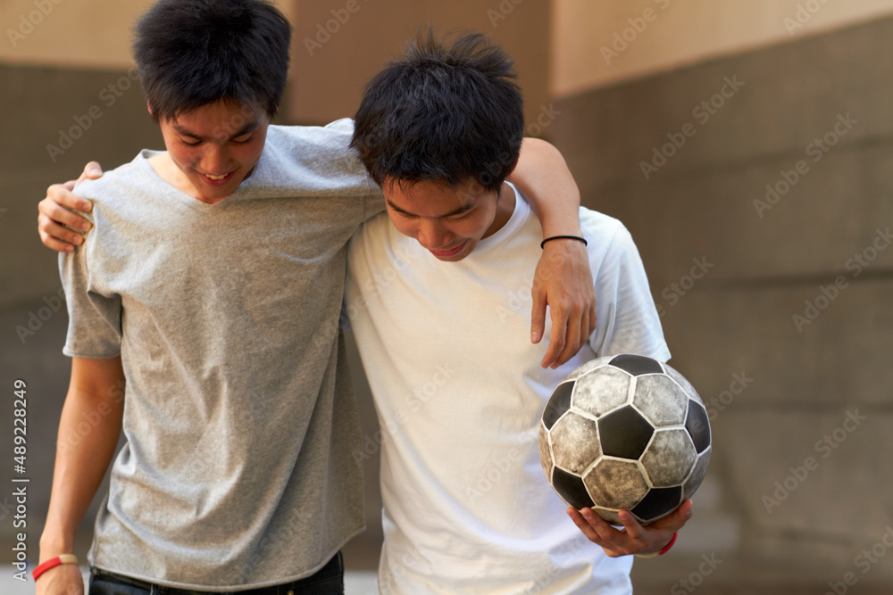 Good game. Two asian boys walking with their arms around eachother holding a soccer ball.