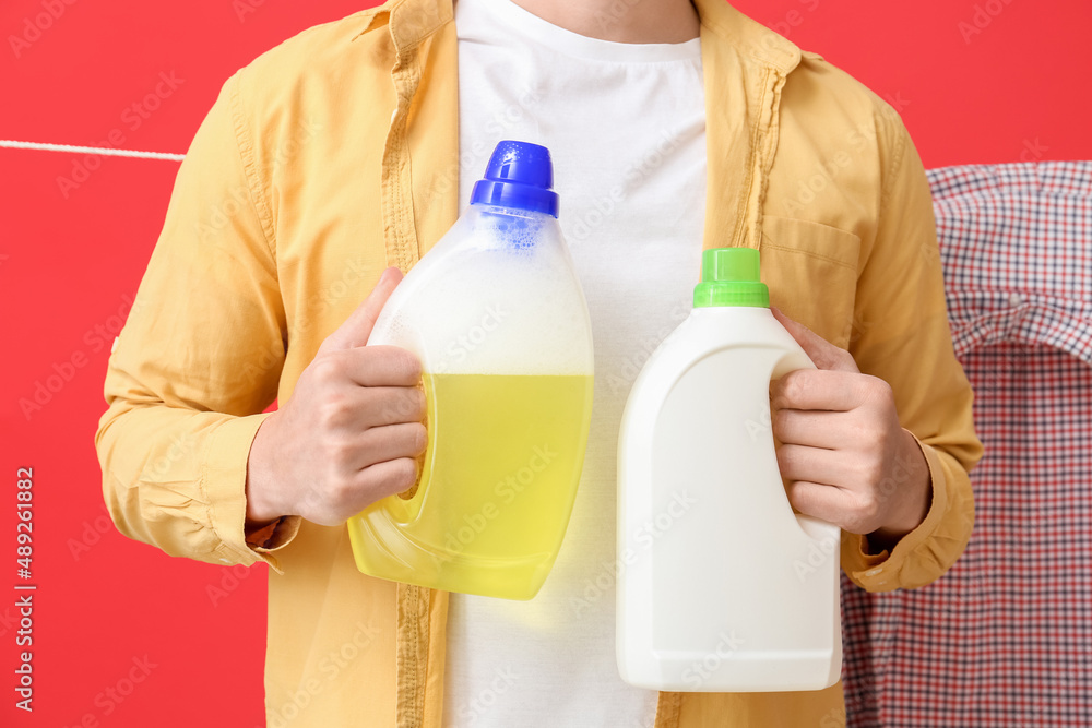 Young man with bottles of detergent and hanging laundry on red background, closeup