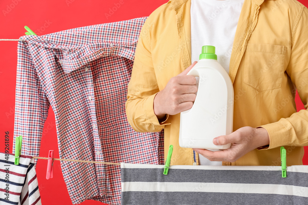 Young man with detergent, hanging laundry and clothespins on red background