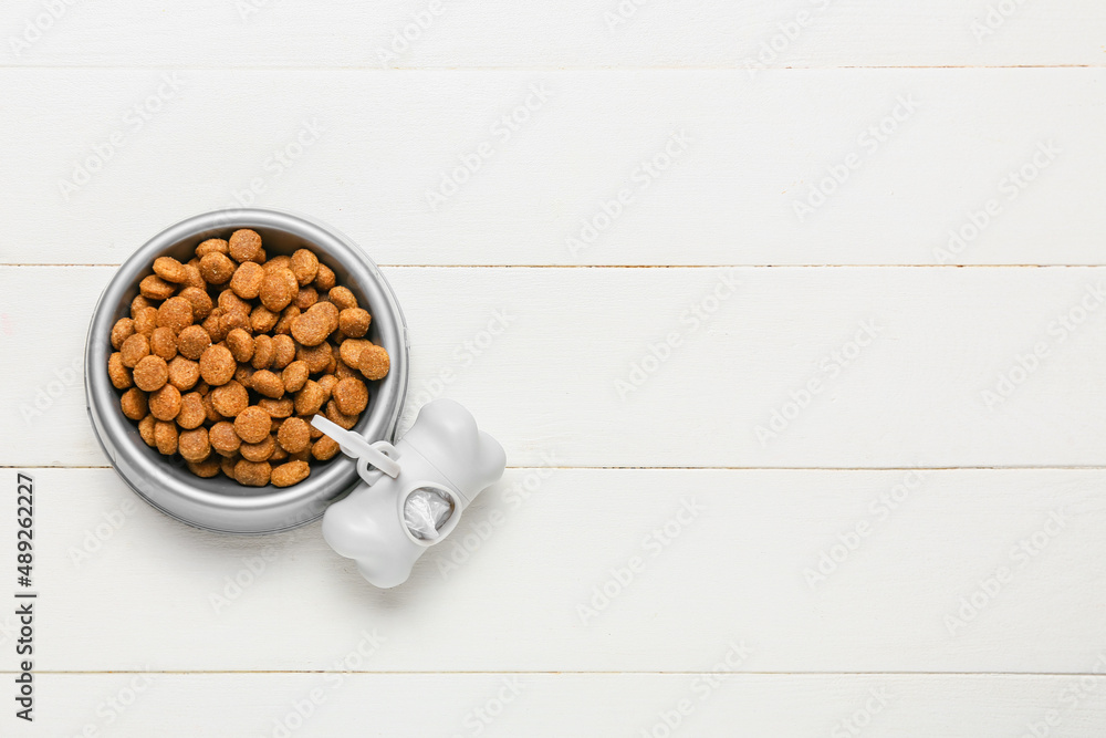 Bowl of dry pet food and waste bags on light wooden background