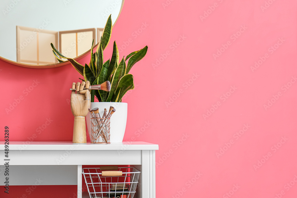 Wooden mannequin hand and holder with makeup brushes on table near pink wall