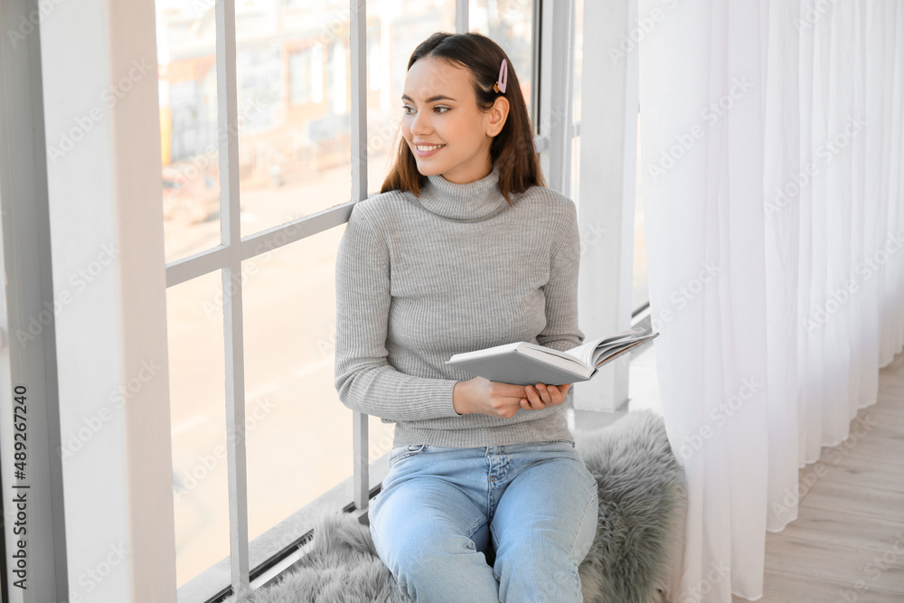 Pretty young woman reading book while sitting on window sill