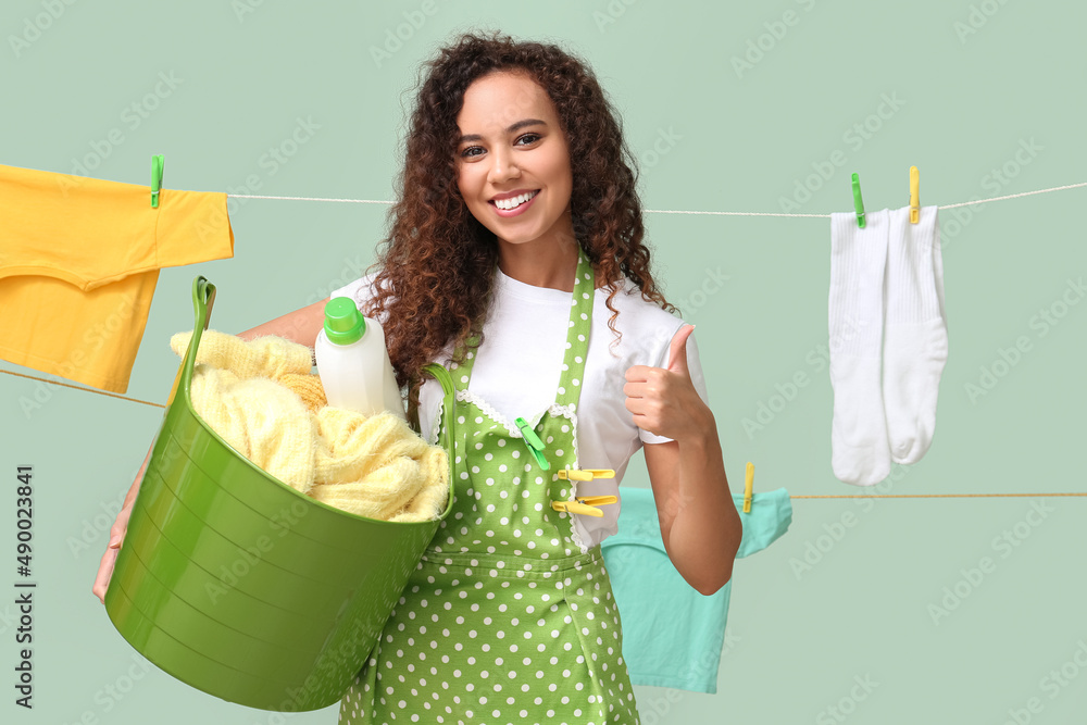 Young African-American woman with laundry basket, detergent and clothespins showing thumb-up on colo