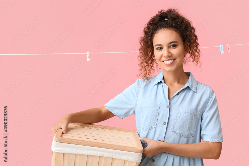 Young African-American woman with laundry basket and hanging clothespins on pink background