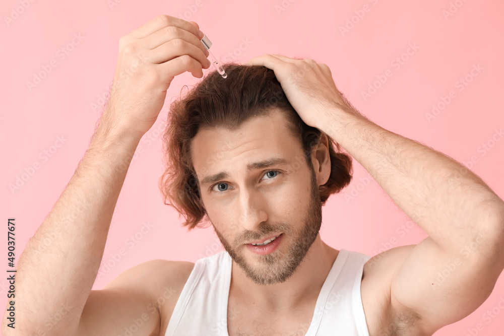 Young man using serum for hair growth on color background