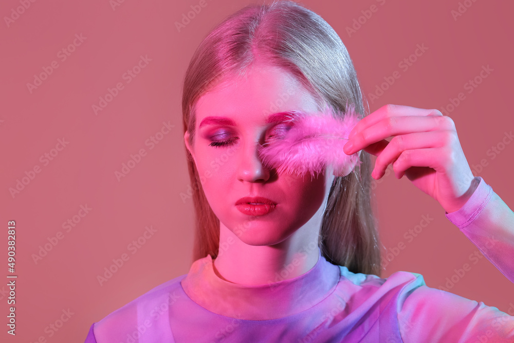 Young woman with creative makeup closing eye with feather on color background