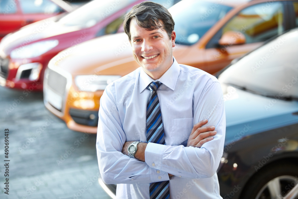 He can give you a great car deal. A man smiling confidently while standing in the lot of his dealers