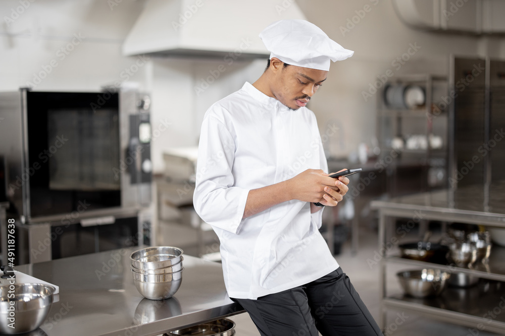 Handsome chef in uniform standing with phone in professional restaurant kitchen. Latin guy using pho