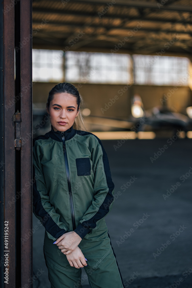 Serious female pilot, leaning onto the airport door, posing for the camera.