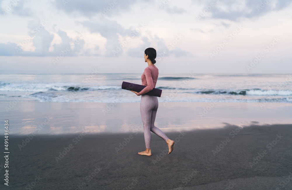 Slim female yogi dressed in trendy sportive wear holding violet mat choosing place for morning train