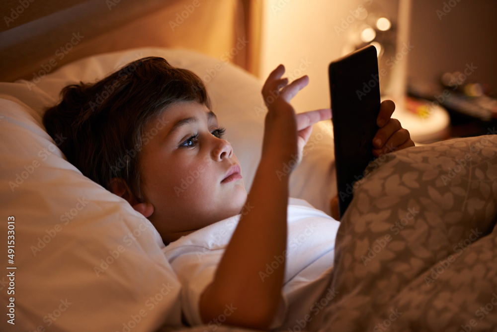 Some light reading before bed. Shot of a young boy playing on a tablet while lying in bed.