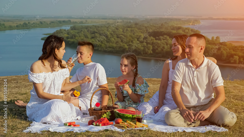 Large happy family in white clothes rests sitting on lush lawn having festive picnic with fresh frui