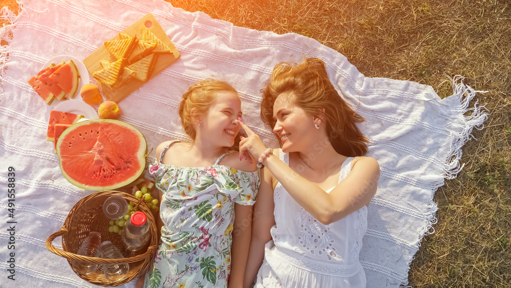 Happy mother touches nose of little daughter lying on white blanket with delicious fruits prepared f