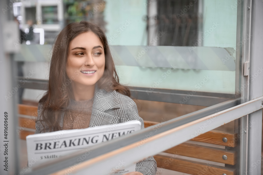 Beautiful young woman with newspaper sitting on bus stop