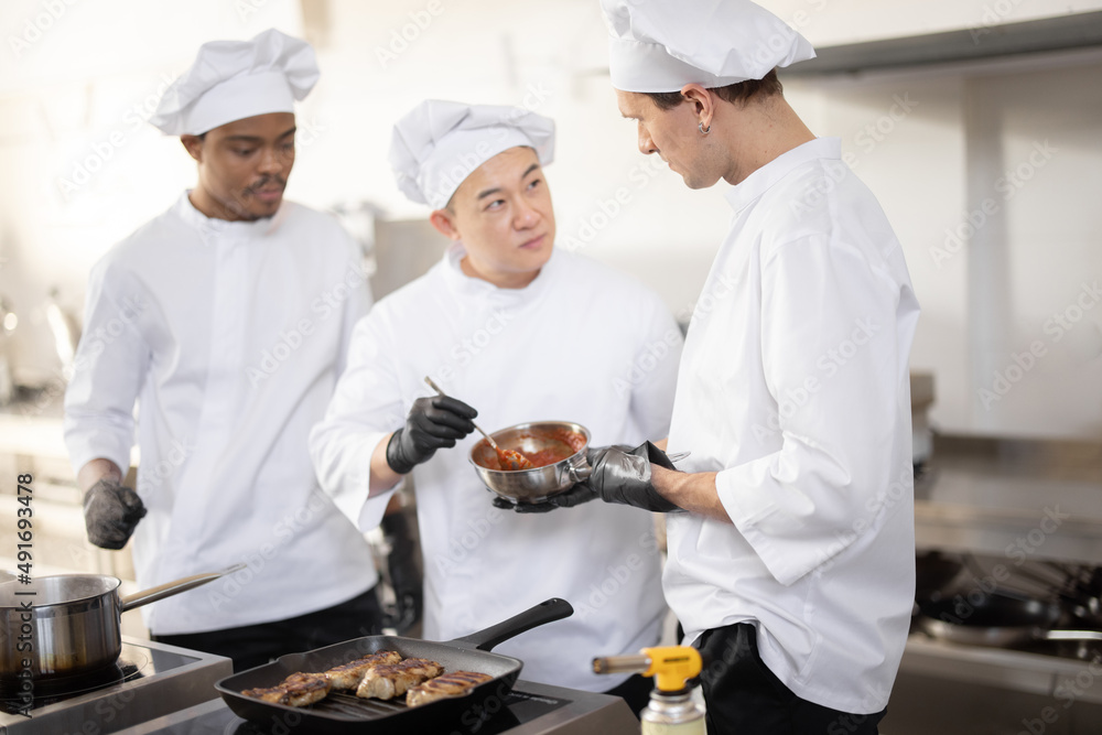 Three chef cooks with different ethnicities tasting sauce with a spoon while cooking in the kitchen.