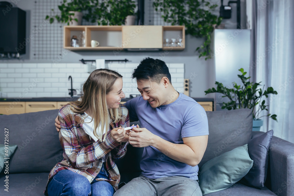 Happy couple of man and woman of different races rejoice in pregnancy test result, young family at h