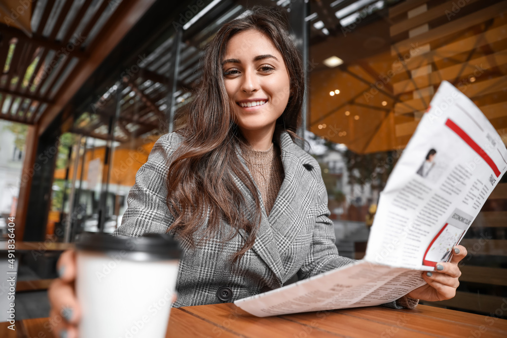 Beautiful young woman with newspaper taking cup of coffee in cafe