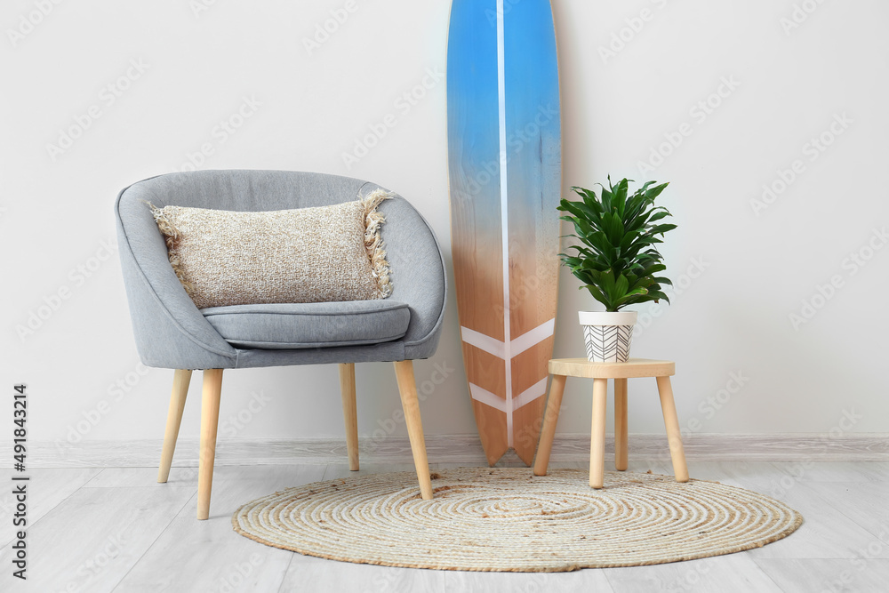 Armchair, stool with houseplant and surfboard near light wall