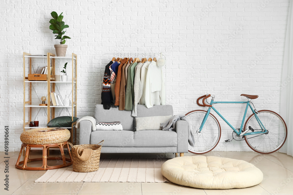 Interior of living room with comfortable sofa and rack with stylish sweaters near white brick wall