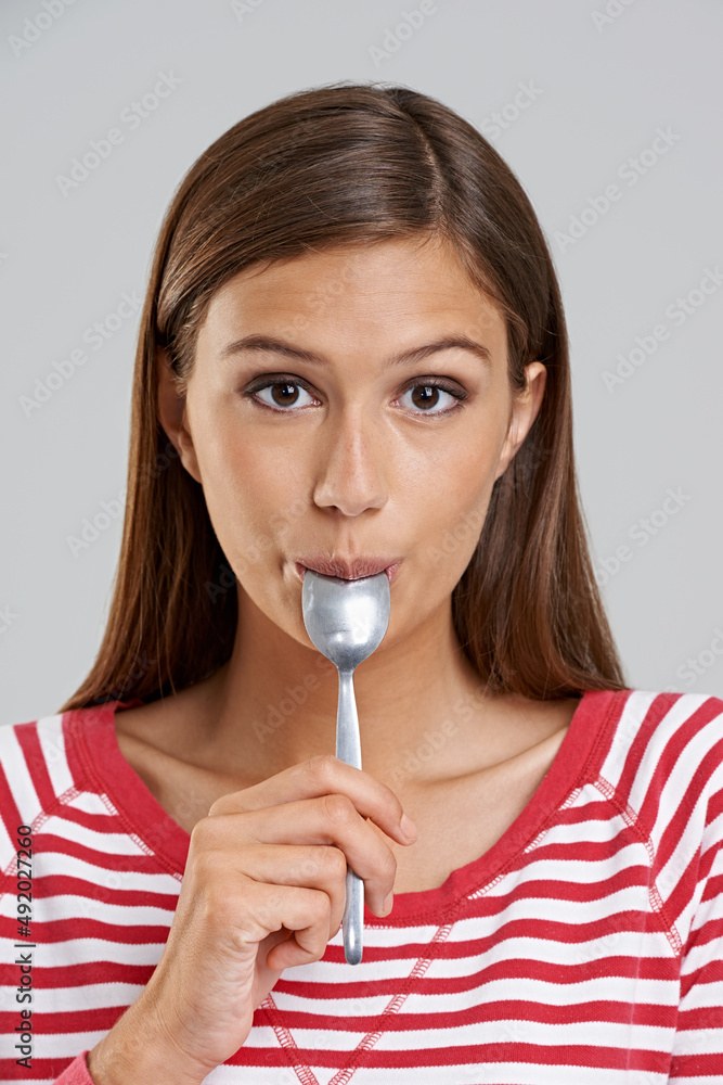 Get every last drop. Studio shot of an attractive young woman with a spoon in her mouth on a grey ba