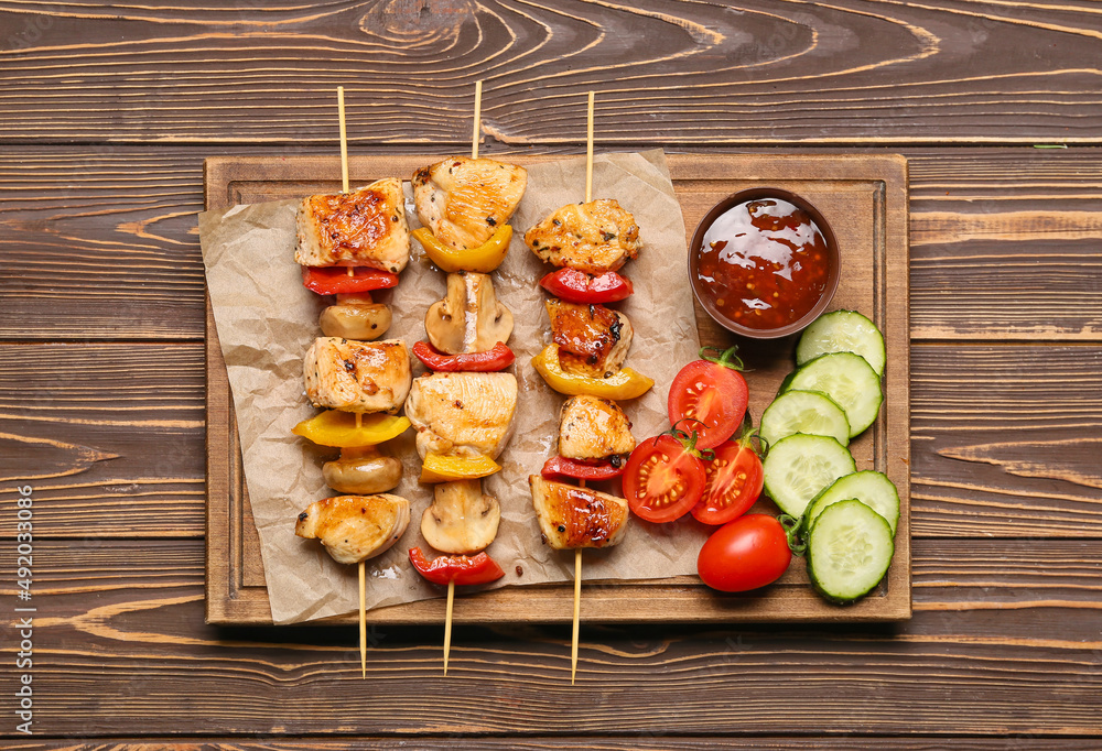 Board of grilled chicken skewers with vegetables and sauce on wooden background