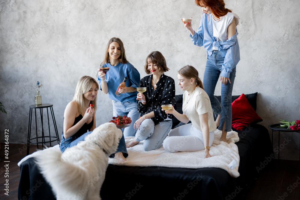 Four young adult girlfriends dressed casual having fun, sitting with drinks and playing with dog on 