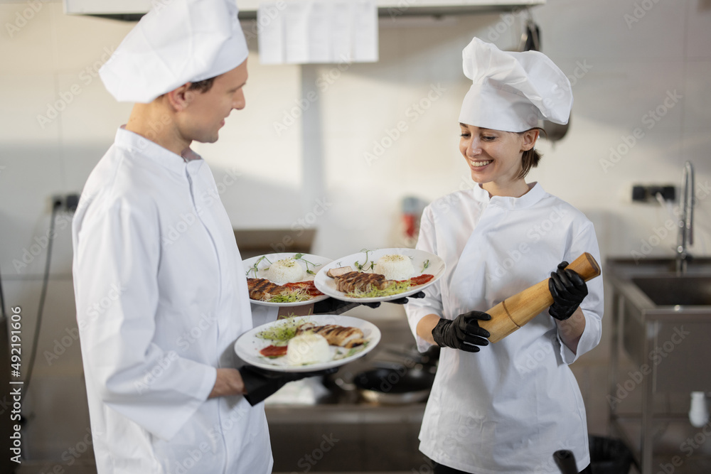 Chef standing with ready meals, talking with female cook in the kitchen. Happy caucasian cook workin