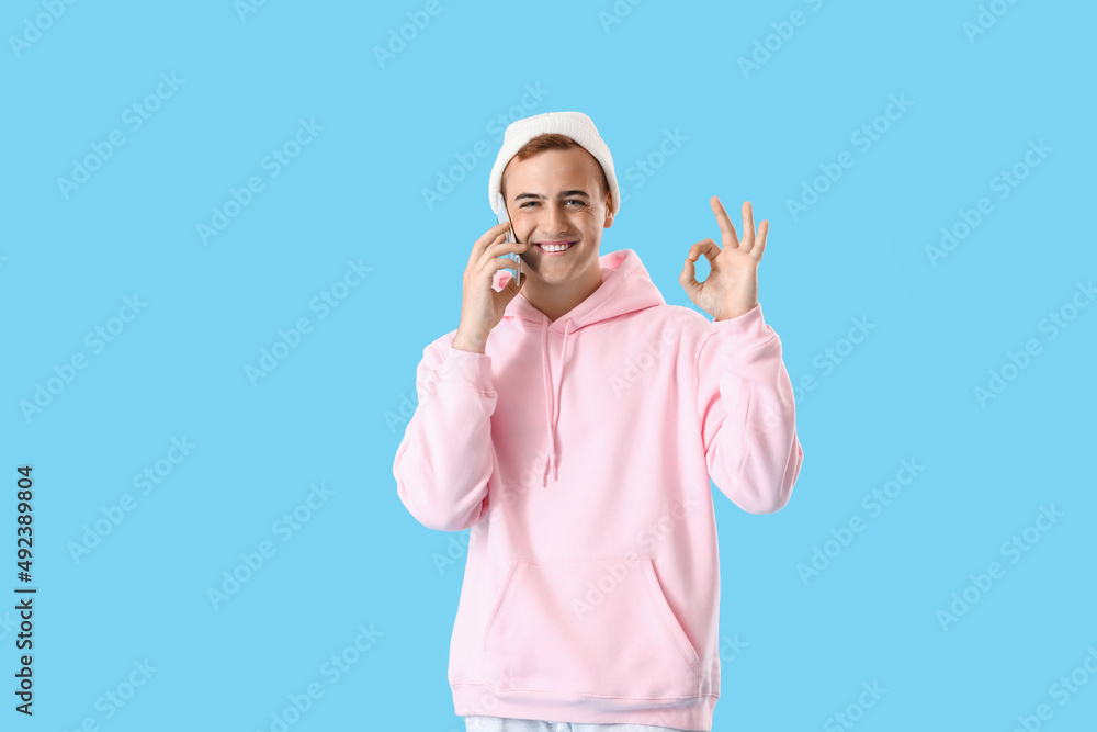 Young guy in stylish hoodie talking by phone and showing OK on color background