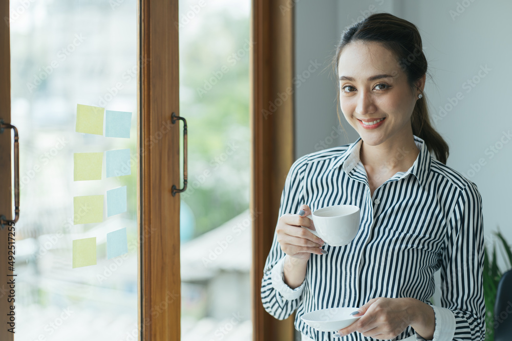 Asian woman working in office with coffee cup, sticky notes on glass wall.