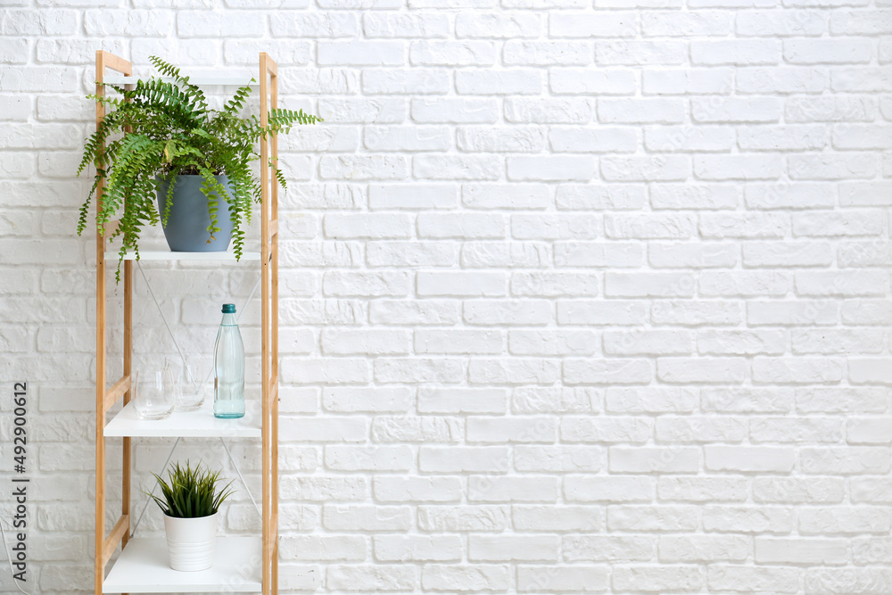 Shelving unit with bottle of water, glasses and houseplants near white brick wall